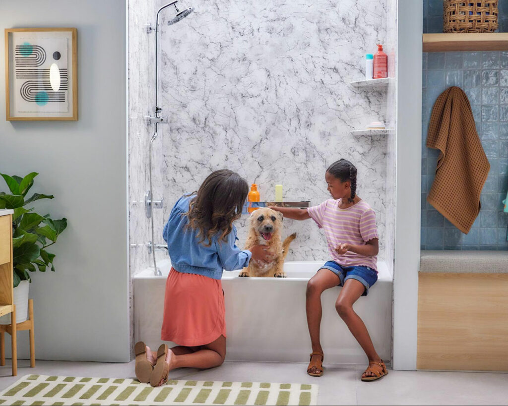 This image depicts a cheerful domestic scene within a bathroom, where a woman and a young girl are giving a bath to a happy golden retriever. The woman, kneeling beside the tub, is gently pouring water over the dog, while the girl, seated on the edge of the tub, is playfully patting the dog. The bathroom has a modern aesthetic with a floor-to-ceiling marble-patterned tub surround and glass doors. Natural light fills the room, highlighting the wooden furniture, a green striped rug, and a hanging wicker basket with a towel. A framed abstract artwork adorns the wall, adding a touch of sophistication to the homely and lively atmosphere.