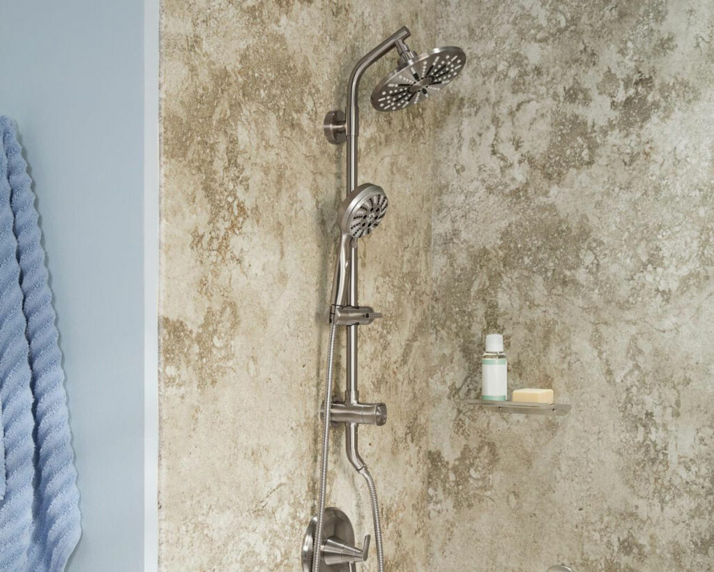 The image shows a close-up view of a modern shower setup against a textured wall with a rustic, stone-like appearance that provides a natural, earthy backdrop. The shower features a shiny chrome dual-function shower system, with both a large overhead rainfall showerhead and a handheld showerhead attached to a vertical bar, allowing for adjustable height. A small glass shelf is mounted to the wall, holding a bottle and a bar of soap, indicating a minimalist approach to bathroom accessories. To the left, the edge of a soft blue towel can be seen, adding a pop of color that contrasts with the neutral tones of the shower area. The focus is on the elegant fixtures and the textured wall, suggesting a blend of luxury and organic design elements.
