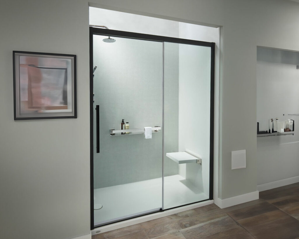 The image presents a modern, minimalistic bathroom shower area. It features a spacious walk-in shower enclosed by a large glass door with a sleek black frame, which complements the shower's modern design. The shower is equipped with a slim rainfall showerhead that matches the black frame of the door. Inside the shower, a simple white bench is attached to the wall, and there's a built-in niche for toiletries. The bathroom's walls are painted in a soft grey, and the floor has wood-effect tiles, enhancing the contemporary feel. On the wall outside the shower, there's a framed abstract painting adding a touch of color to the neutral palette. To the right, the reflection of a well-lit bathroom vanity with various bottles can be seen in a large mirror, contributing to the clean and serene atmosphere of the space.
