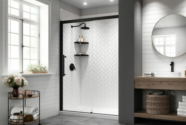 A modern bathroom featuring a herringbone pattern of textured white tiles on the shower wall. The room has matte black shower fixtures and a large, round mirror with a thin, gold frame. There's a dark grey sliding glass door for the shower, and the floor is laid with large, slate grey tiles. A wooden vanity with a white countertop holds a rectangular sink with a black faucet. A small, black, metal-framed trolley holds towels and toiletries, complementing the room's modern chic aesthetic. A large window with white trim lets in natural light, enhancing the clean and fresh ambiance of the space.